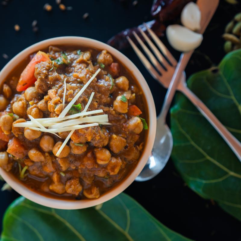 A bowl of spicy chickpea curry garnished with julienned ginger on a dark background. Leaves, a fork, and garlic cloves are placed nearby.