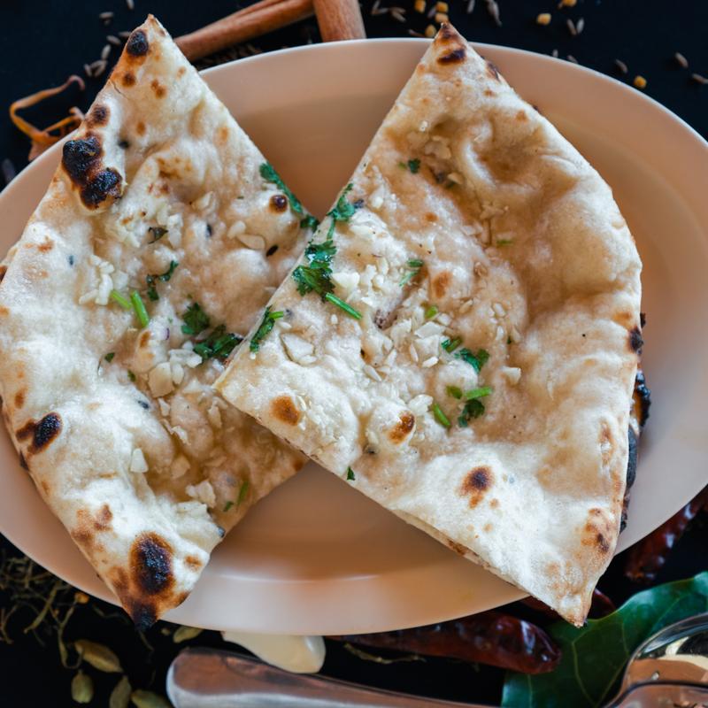 Close-up of a plate with two pieces of golden-brown garlic naan topped with cilantro. The background includes spices, adding to an aromatic ambiance.