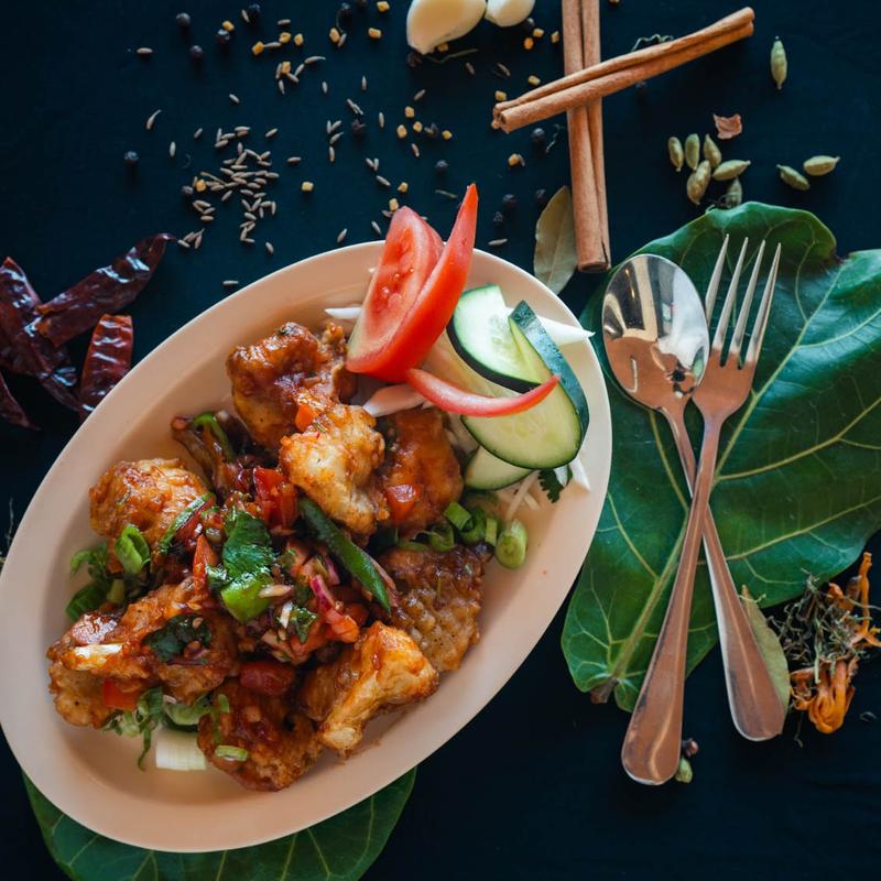 A plate of crispy fried chicken garnished with cucumber and tomato slices on a green leaf. Surrounded by spices, garlic, and utensils on a dark surface.
