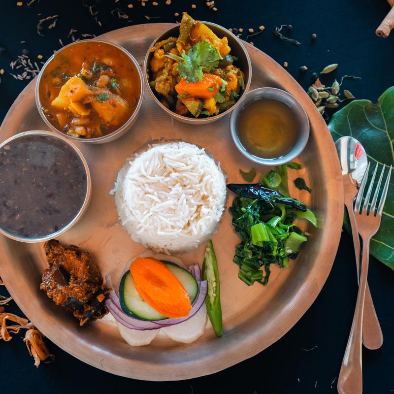 A traditional meal on a metal plate featuring rice, lentils, vegetables, yogurt, pickles, and greens on the side. Fork and spoon placed nearby.