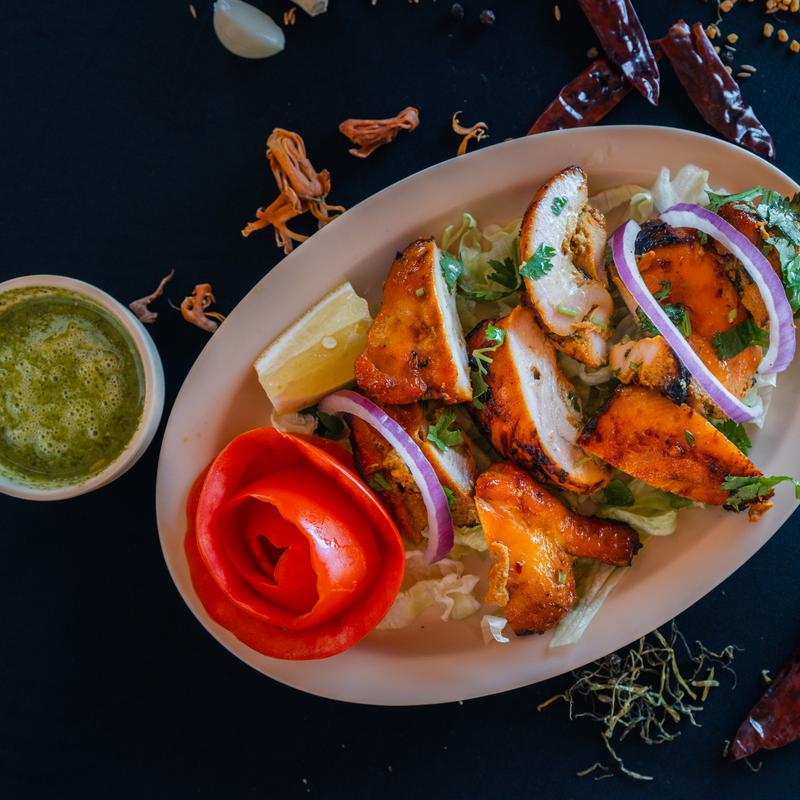 A white plate with grilled chicken slices topped with cilantro and red onion on lettuce. Beside it, a tomato rose and lemon wedge. A bowl of green sauce is nearby.