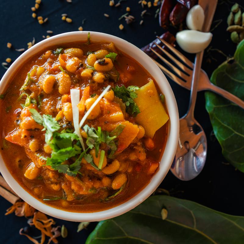 A colorful bowl of spicy curry with black-eyed peas, garnished with cilantro and sliced ginger. Surrounded by garlic, utensils, and spices on dark leaves.