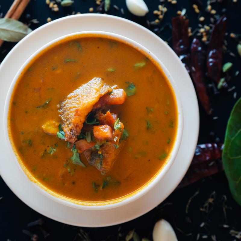 A bowl of spicy fish curry garnished with fresh herbs. The rich, orange-red broth contains chunks of fish and vegetables, surrounded by spices.