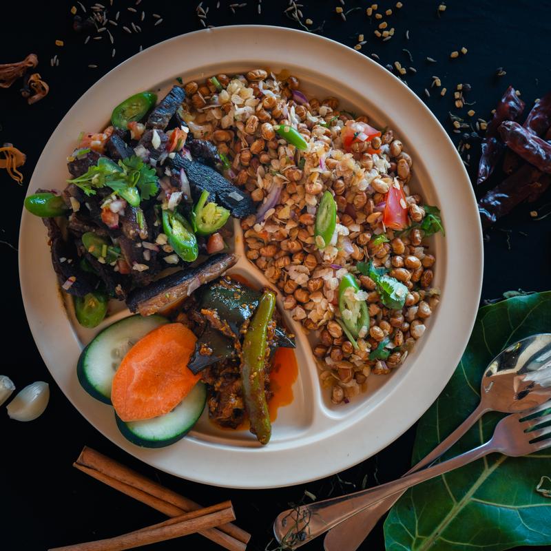 A divided plate featuring spicy stir-fried beef salad, chickpeas with peppers and tomatoes, and pickled vegetables. Surrounded by spices and herbs.