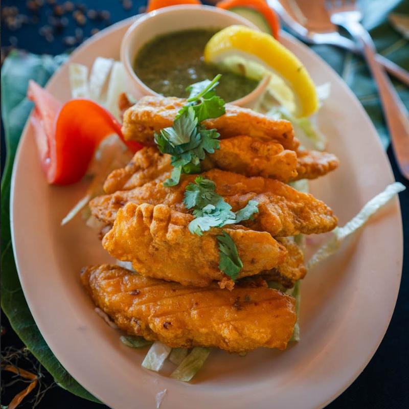 Crispy fried fish fillets on a plate garnished with cilantro, served with green chutney, lemon wedge, and sliced tomatoes, creating a fresh and appetizing feel.