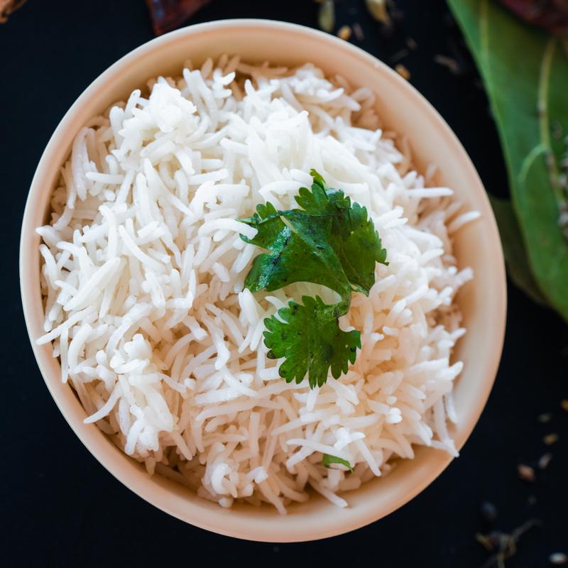 A bowl of fluffy white rice garnished with a sprig of fresh cilantro. The image conveys a sense of freshness and simplicity against a dark backdrop.