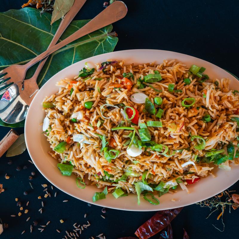 A plate of colorful fried rice garnished with chopped green onions and vegetables on a dark surface. Nearby are a fork, spoon, and some spices.