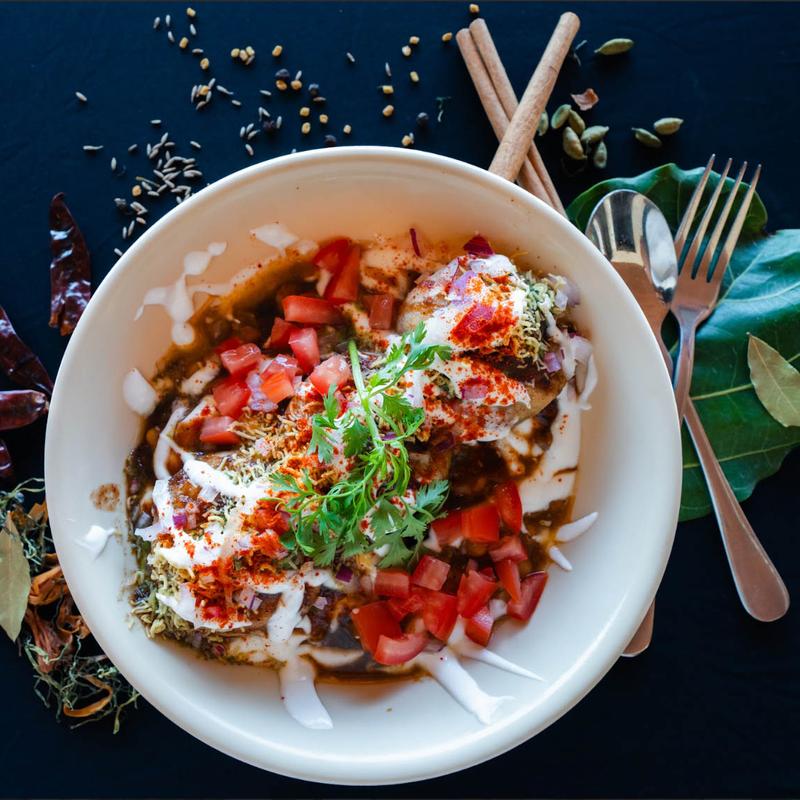 A bowl of vibrant Indian chaat topped with yogurt, chopped tomatoes, spices, and herbs. Surrounded by various spices, cinnamon sticks, and cutlery.