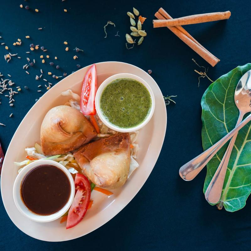 Oval plate with two golden-brown samosas surrounded by sliced tomatoes, cabbage, and two dips, one green and one brown, on a dark tablecloth.
