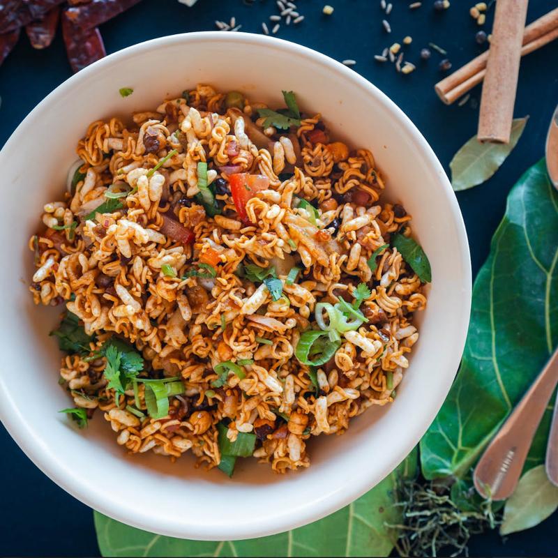 A bowl of spicy noodle salad with fresh herbs, diced vegetables, and puffed rice on a leaf, surrounded by spices and wooden cutlery on a dark surface.
