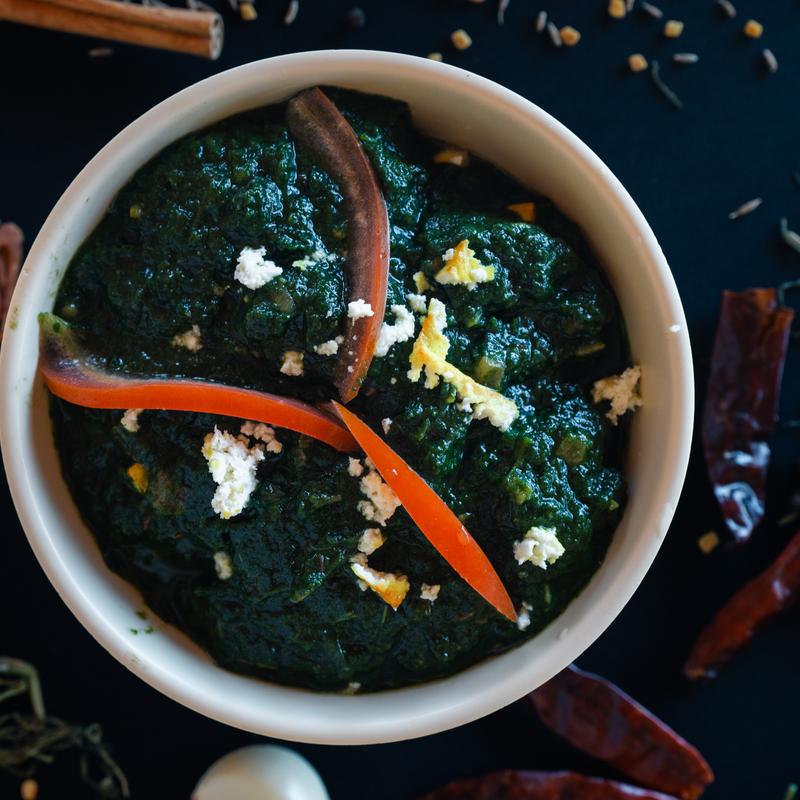 A bowl filled with vibrant green curry, topped with small white cheese crumbles and garnished with red chili slices. Surrounded by whole spices creating a rich, aromatic atmosphere.