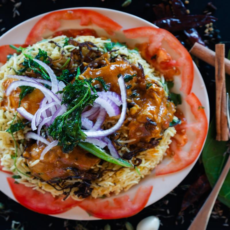 A vibrant dish of yellow rice topped with chicken curry, garnished with purple onion slices and herbs, and surrounded by tomato slices, evokes a savory and colorful appeal.