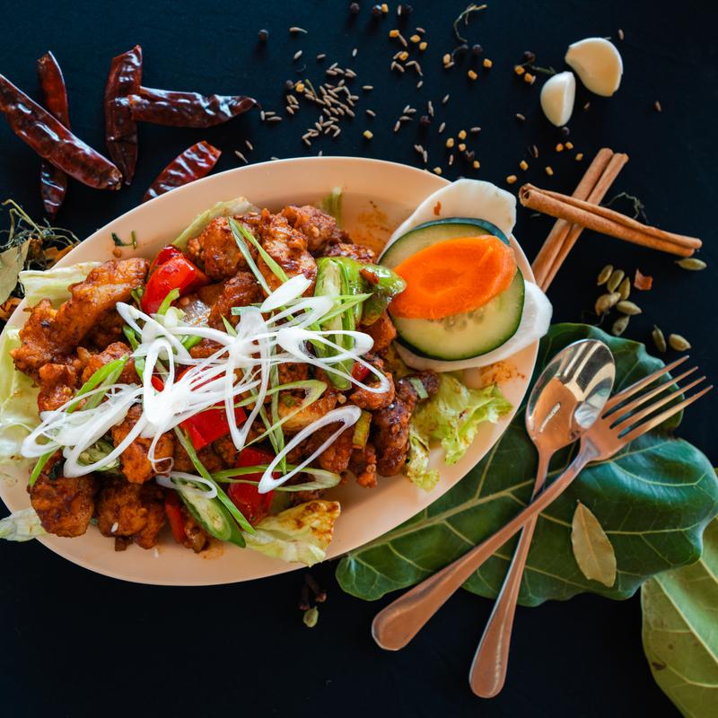 Plate of spicy stir-fry with vegetables, garnished with green onions, surrounded by spices and herbs on a dark table with a fork and spoon.