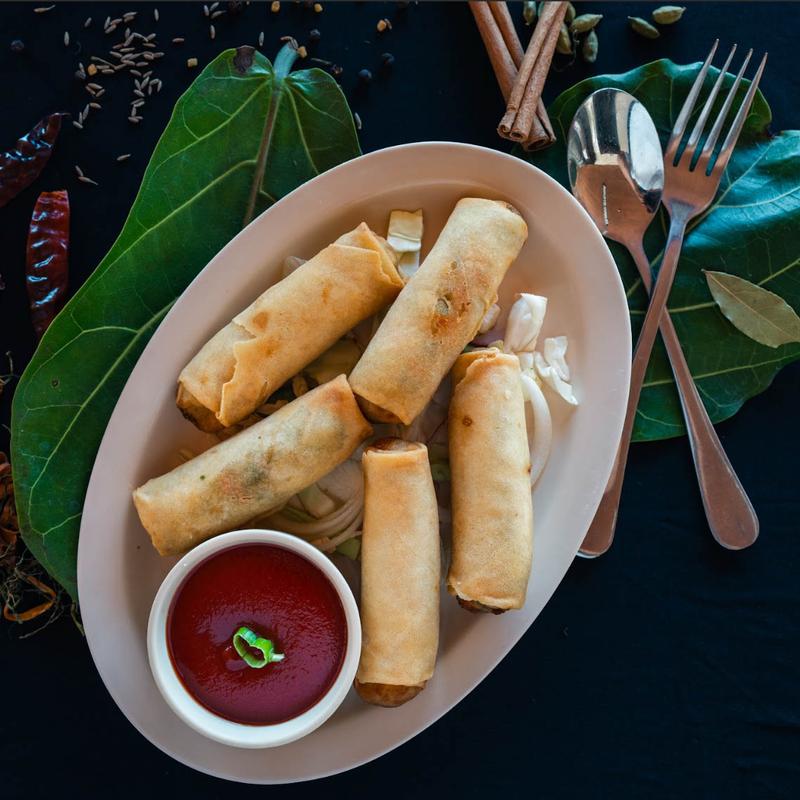 Five golden spring rolls on a white plate with a small bowl of red sauce, garnished with a green chili slice. Surrounding are large green leaves, spices, and cutlery, creating an elegant, appetizing display.