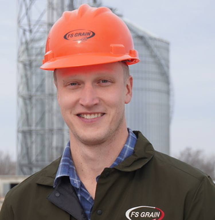 Man wearing an orange FS Grain hard hat and dark jacket with FS Grain logo, standing outdoors with grain silos in the background.