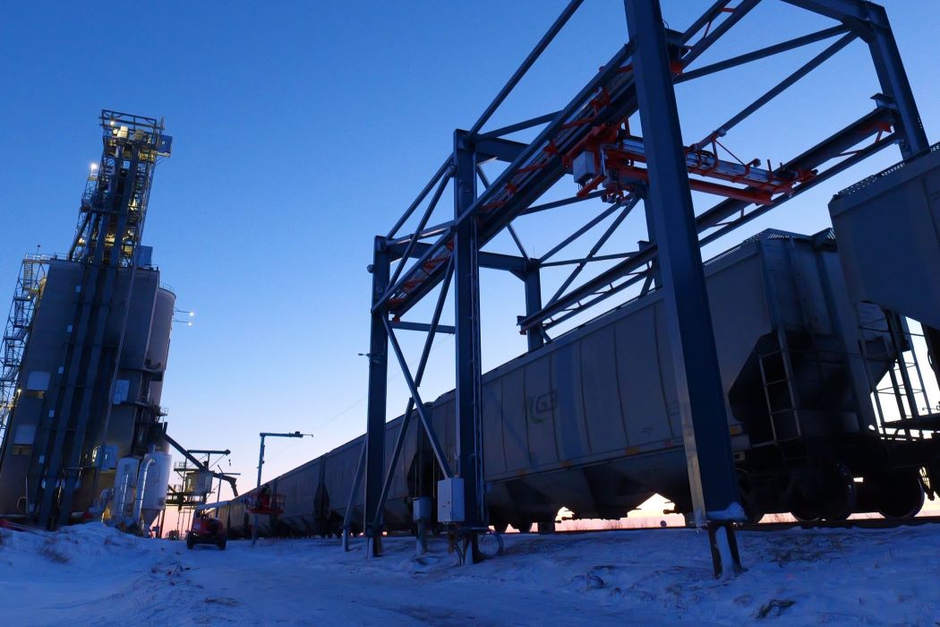 Industrial train cars under a metal loading structure next to a tall facility tower at dusk with snow-covered ground.