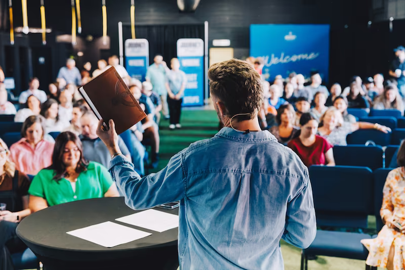 Person holding up a book while speaking to an audience seated in rows in a conference or lecture setting.