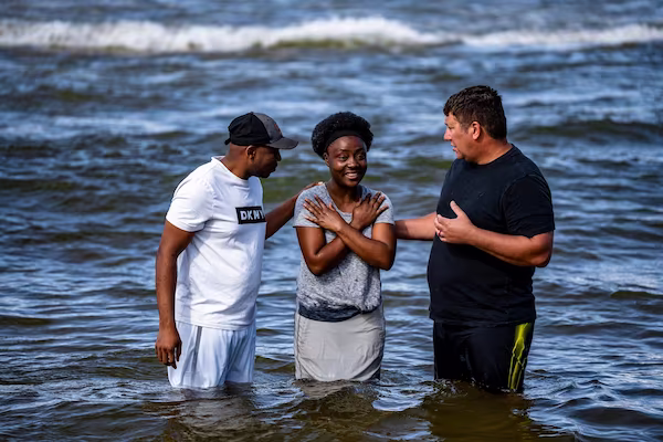 Three adults standing waist-deep in water at a beach, with one woman in the middle smiling and two men on either side talking to her.