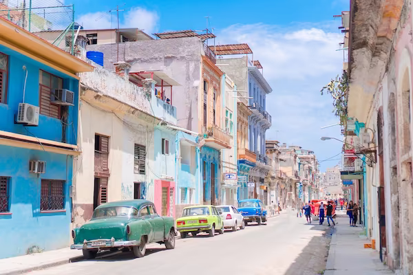 Sunny street lined with colorful old buildings and vintage cars parked along the side with people walking in the distance.