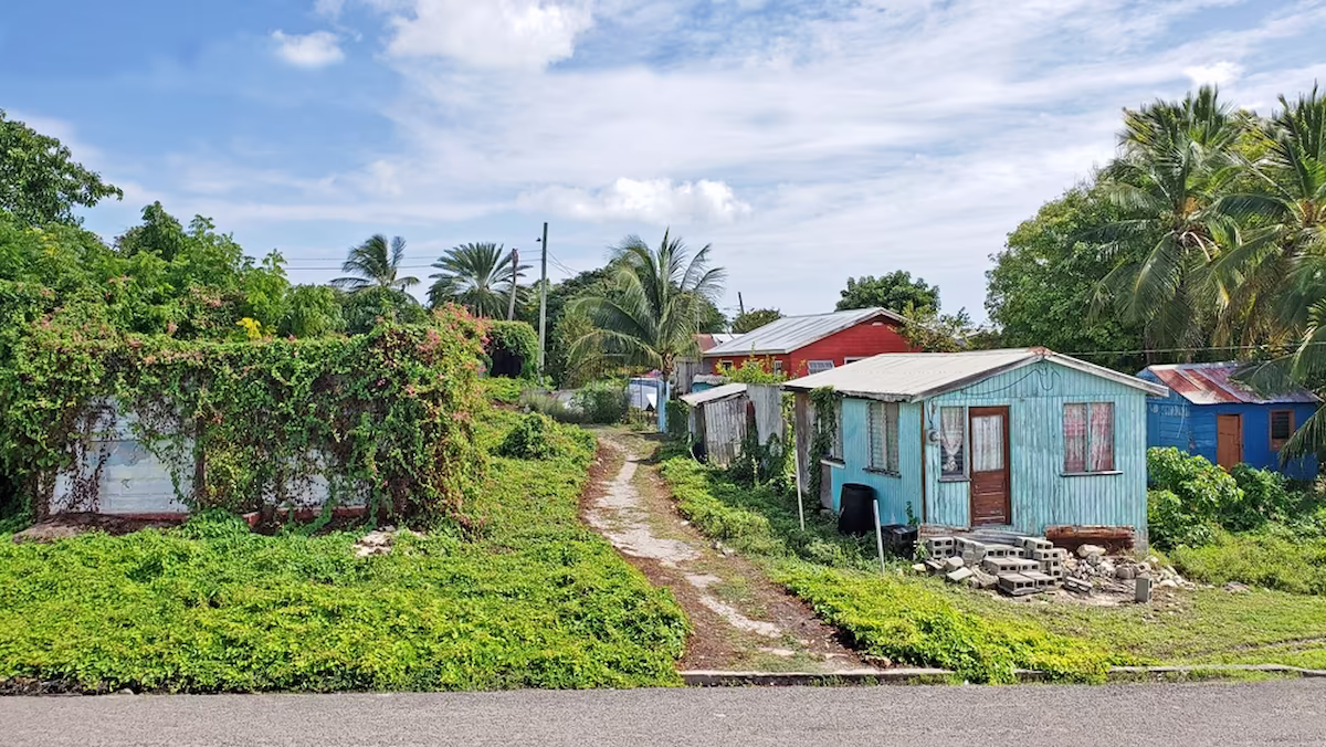 Dirt path flanked by small colorful houses surrounded by lush greenery and palm trees under a partly cloudy sky.