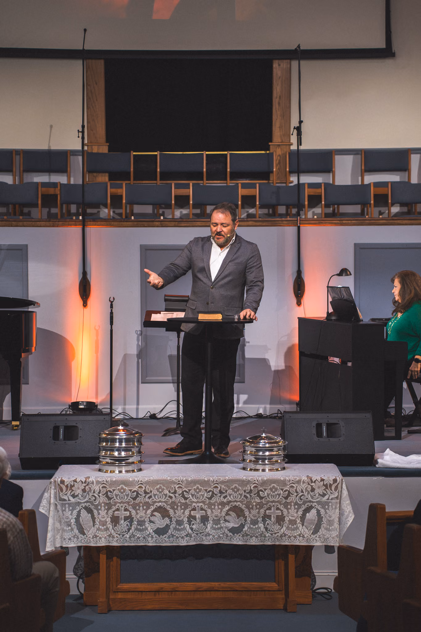 Man speaking at church podium with communion trays on lace-covered table in foreground.