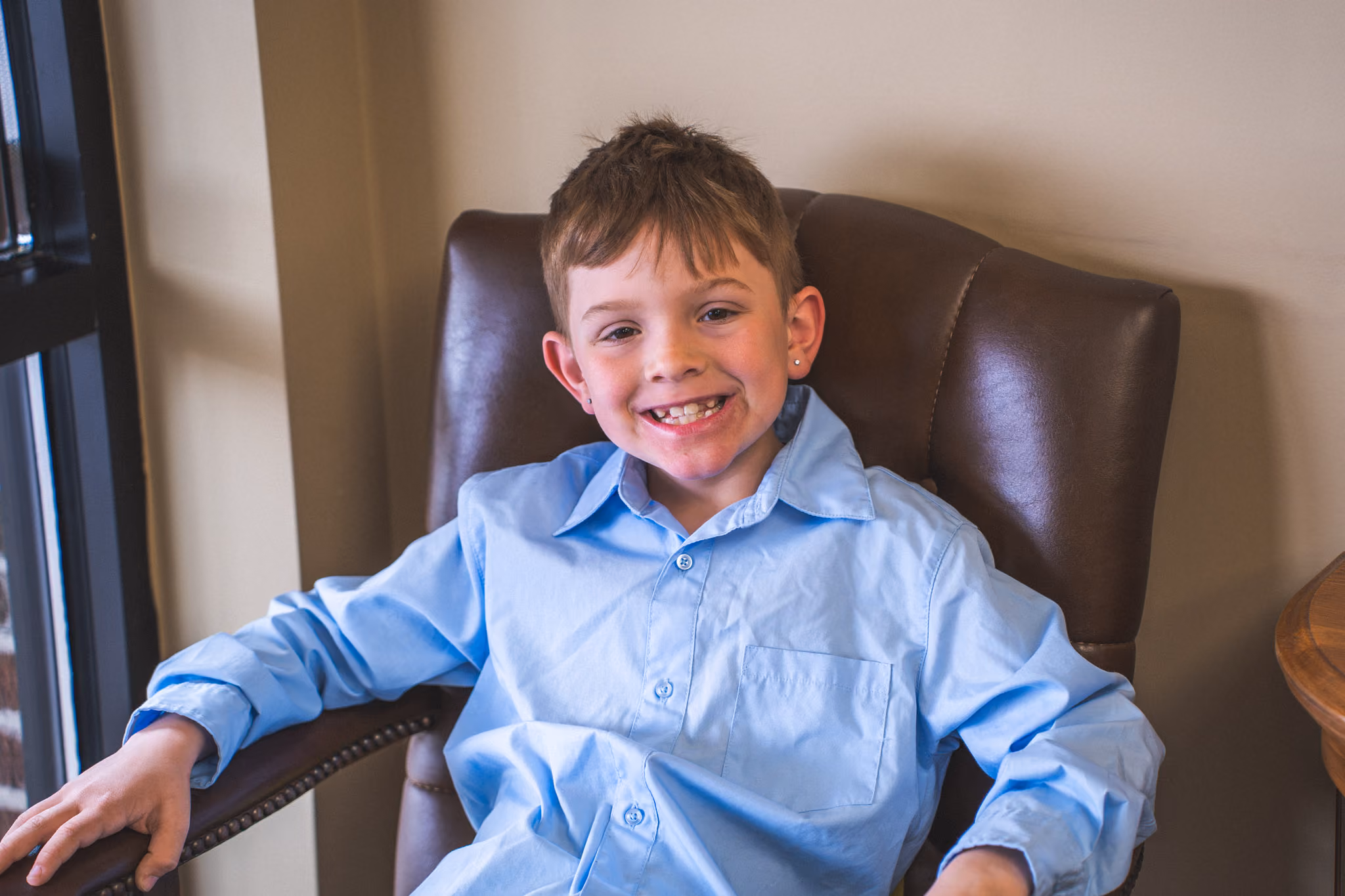 Smiling young boy in a blue button-up shirt sitting in a brown leather chair near a window.