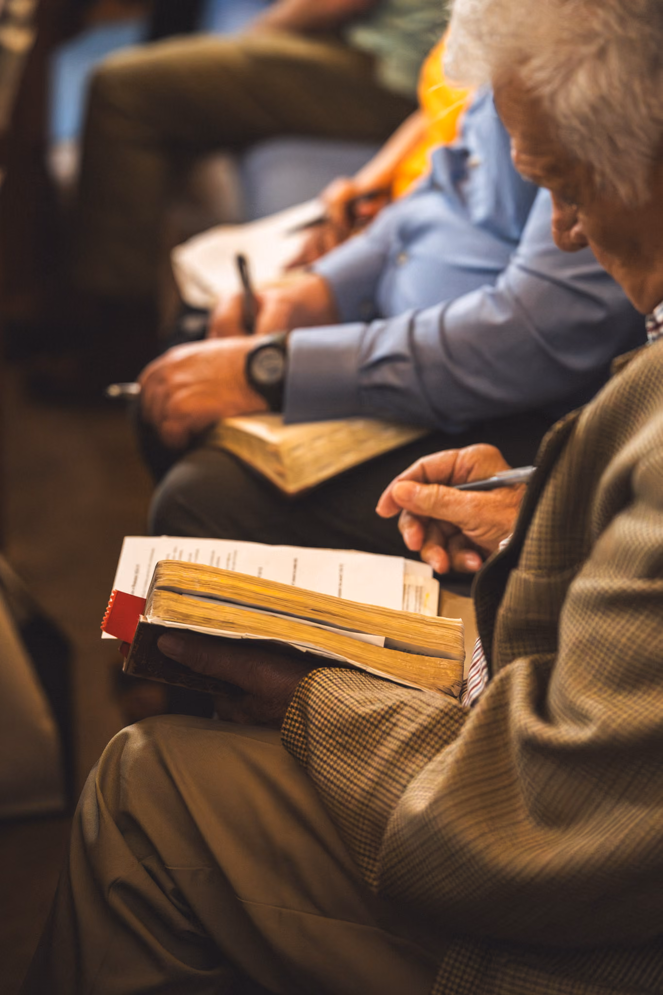 Elderly people seated, reading and writing in books or Bibles during a group gathering.