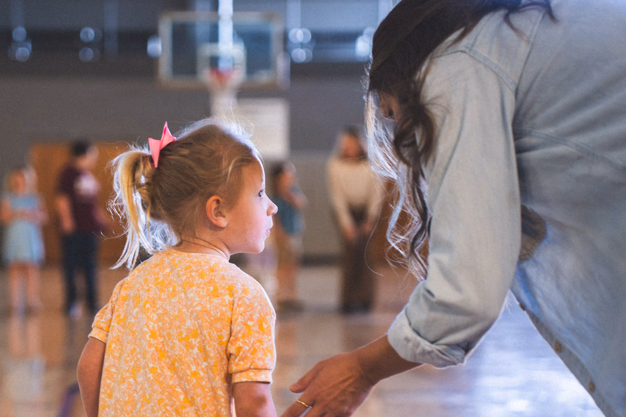 Young girl with pink bow talking to woman in a gym with blurred background figures.