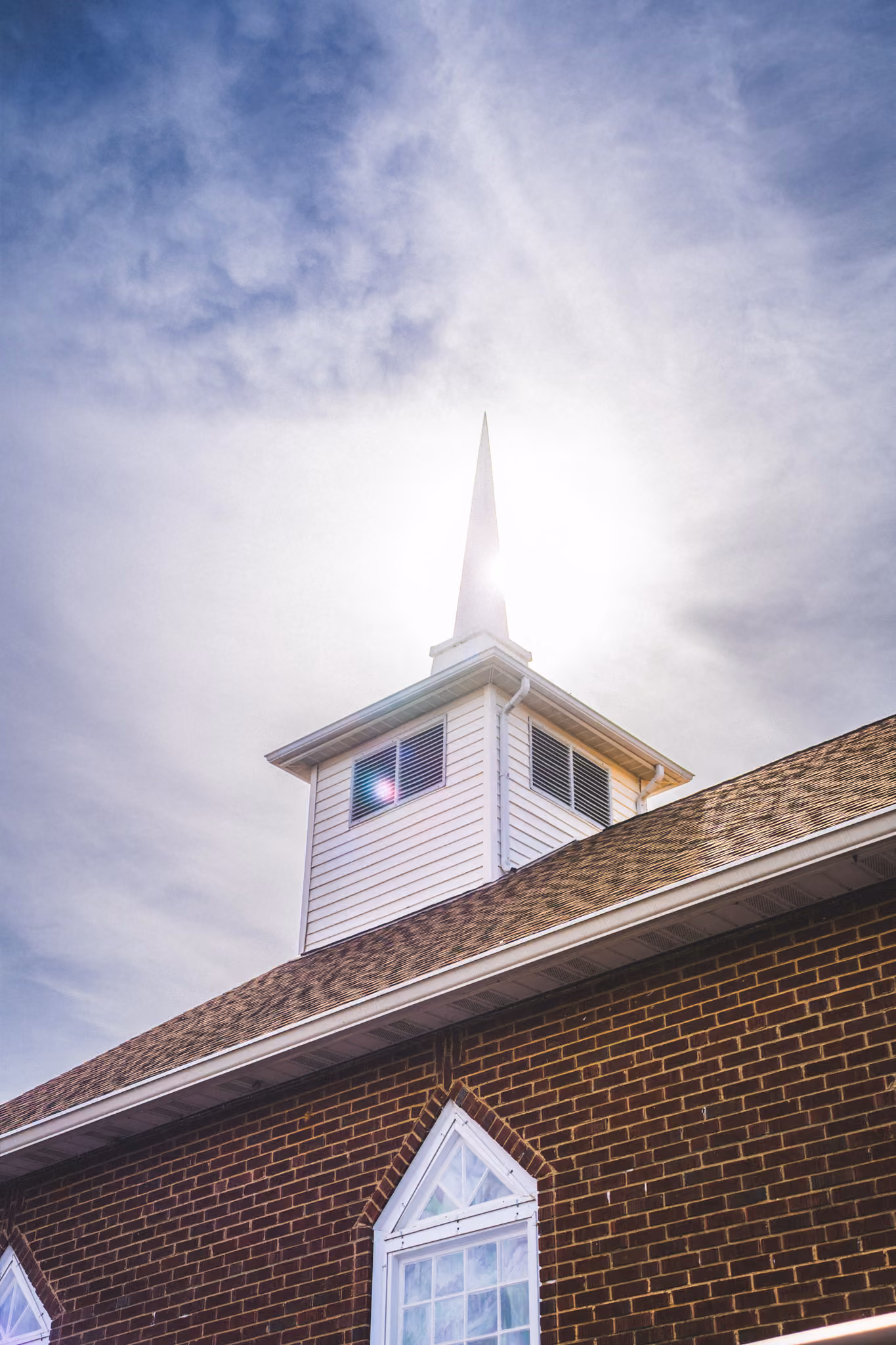 Sun shining behind the steeple of a brick church with white siding and windows.