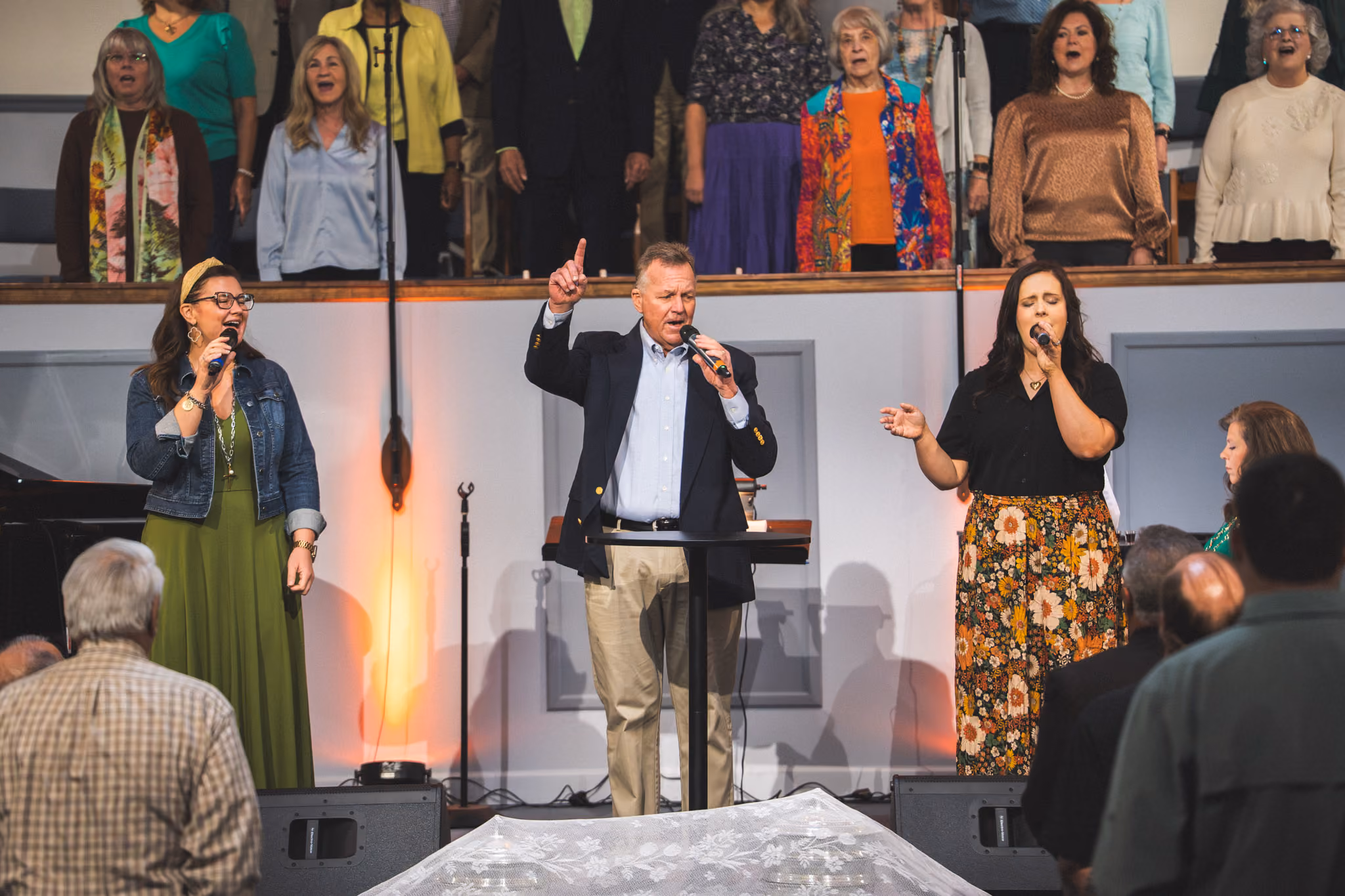 A man and two women singing with microphones on a church stage as a choir sings behind them.