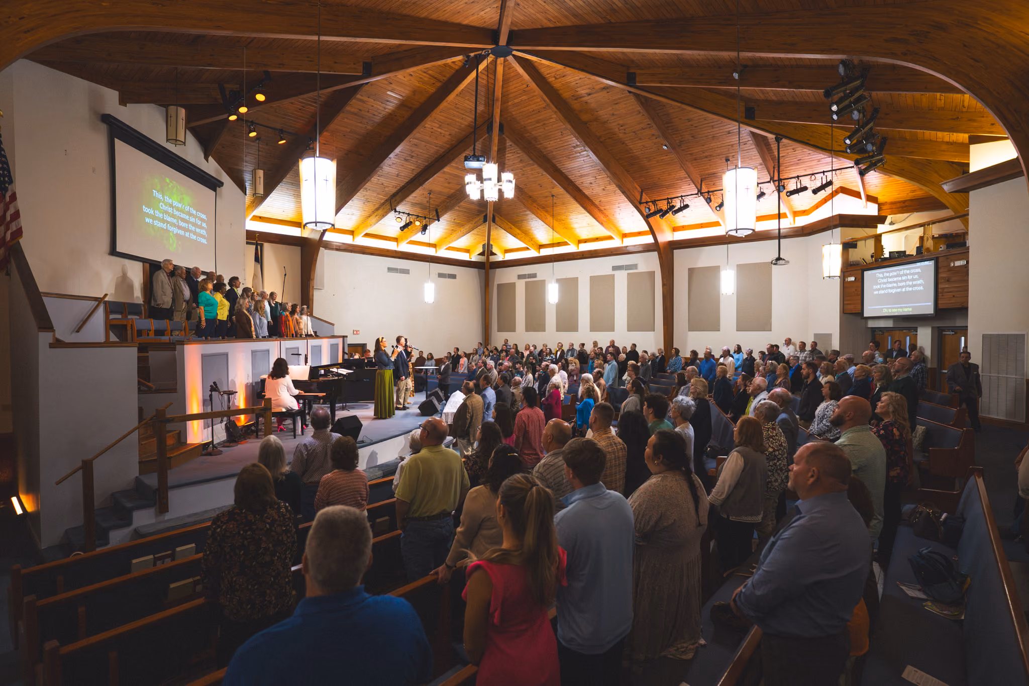 Congregation standing and facing choir and musicians performing in a wooden church sanctuary.