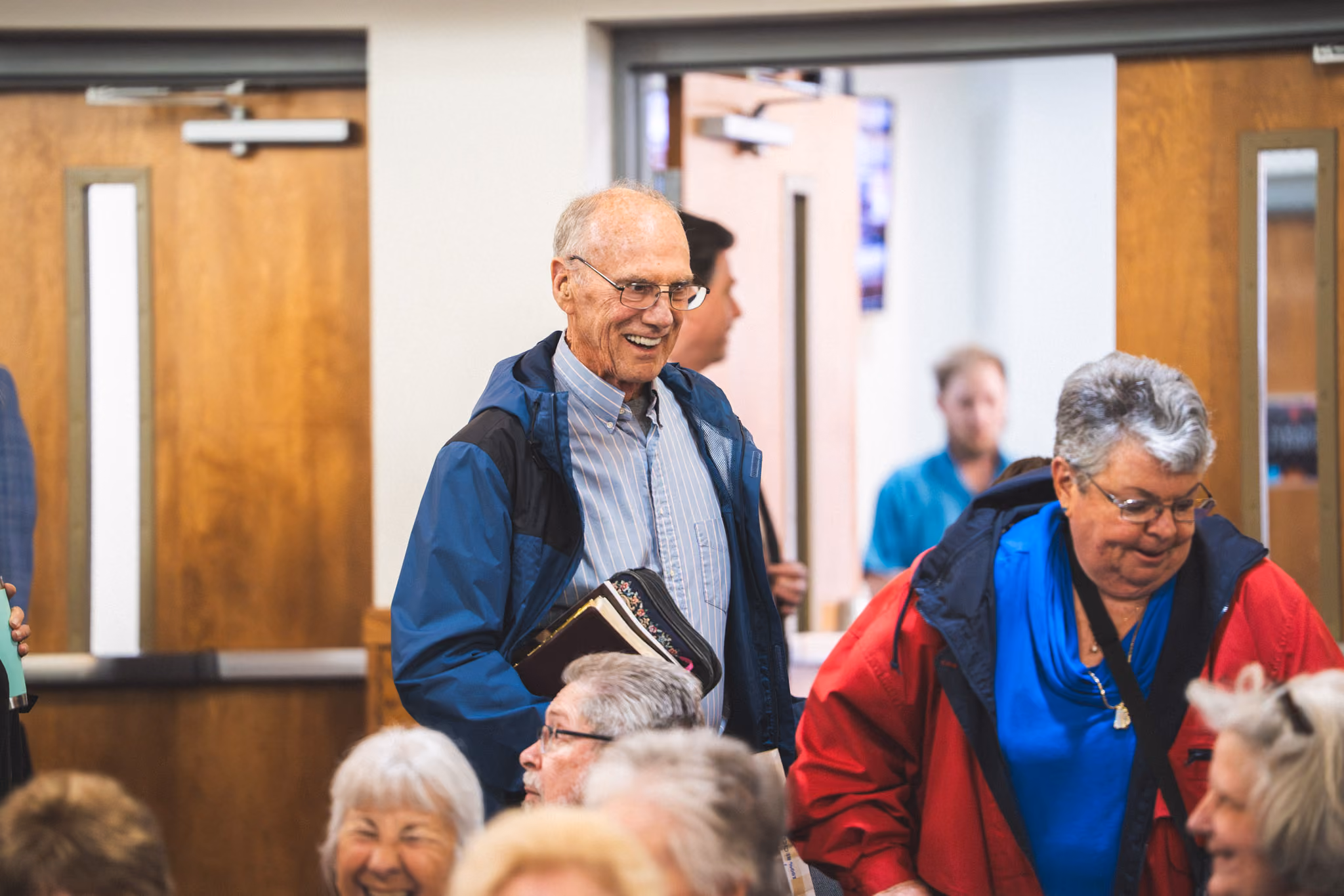 Elderly man smiling and holding a Bible inside a room with other engaged people.