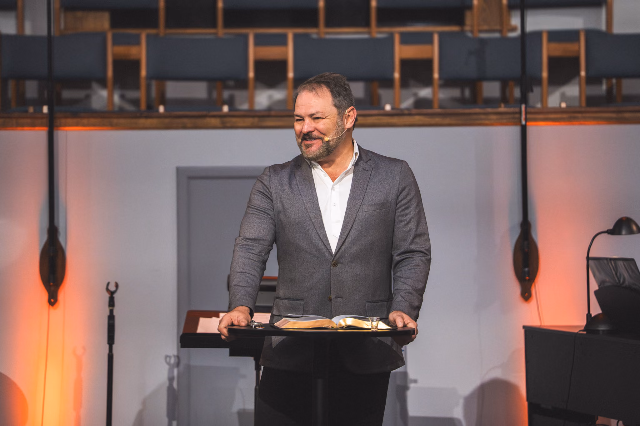 Man in gray suit standing behind a lectern with an open book, speaking with a microphone headset.