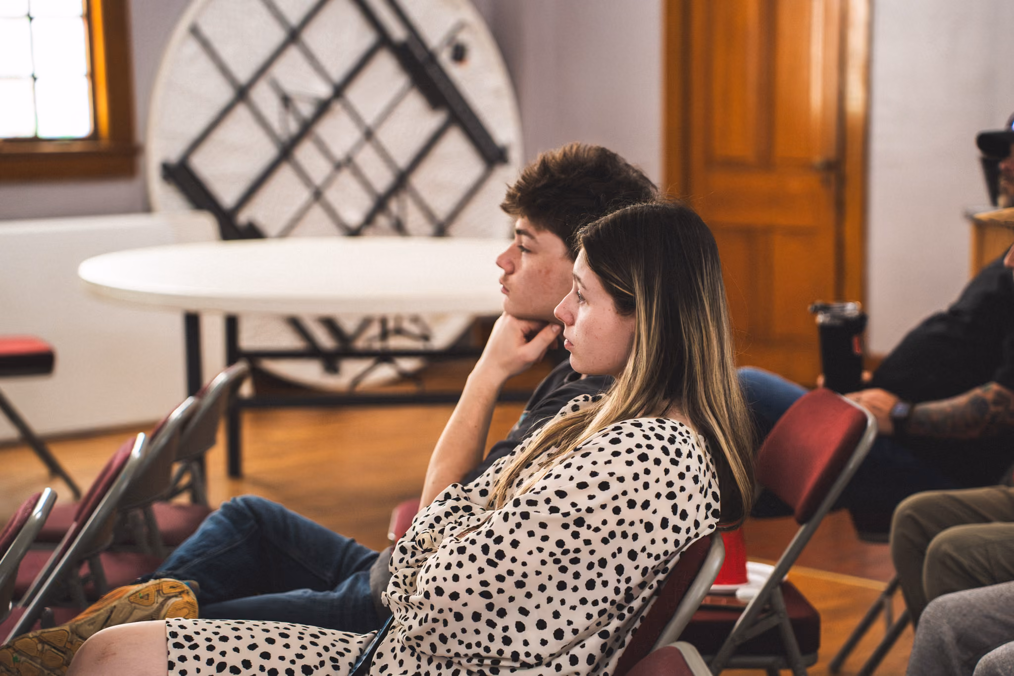 Two young people sitting on red chairs attentively listening in a room with wooden floor and door.