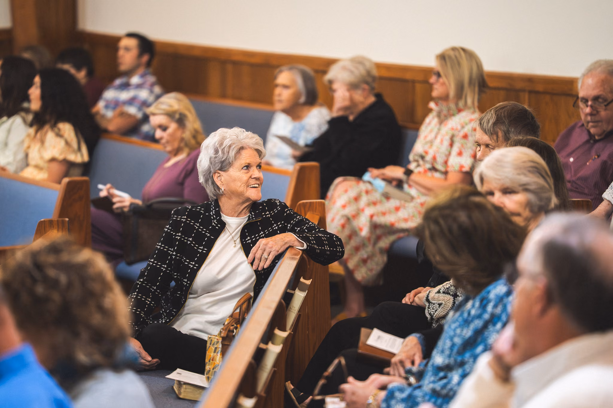 People seated in church pews, an elderly woman in front smiles and talks with others nearby.