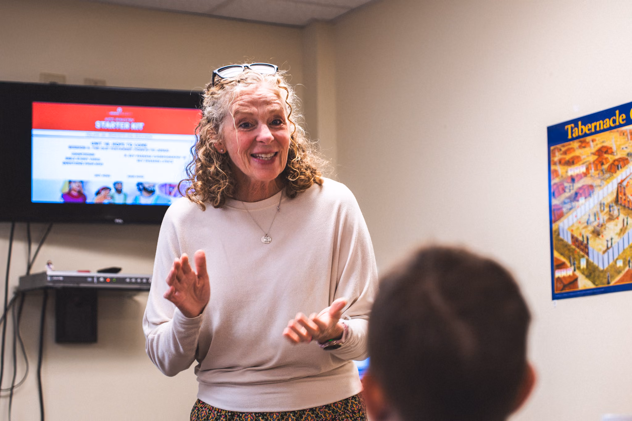 Woman with curly hair and glasses on head speaking to a child in a classroom with a screen and poster.