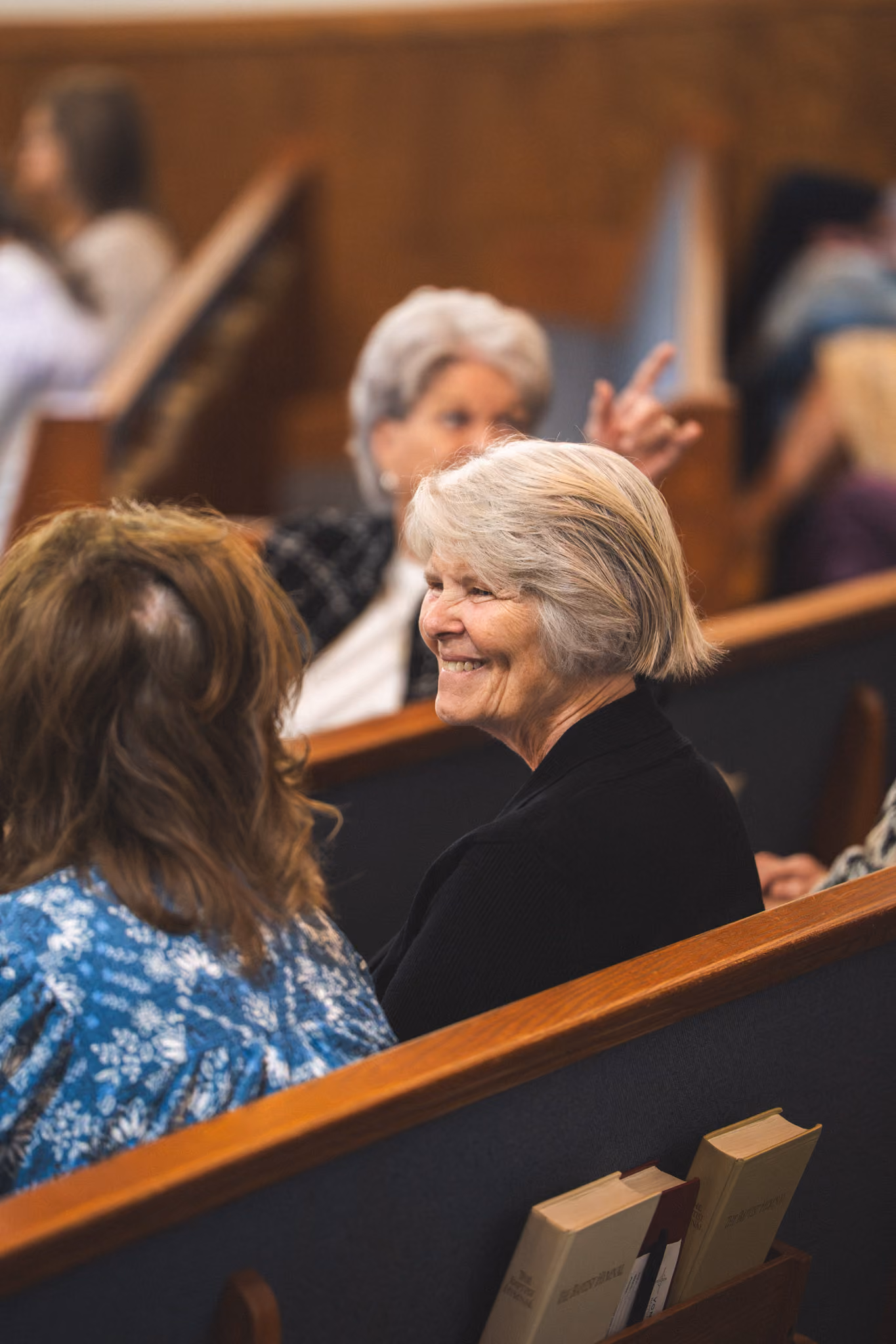 Two women sitting and smiling in church pews with hymnals in the rack behind them.