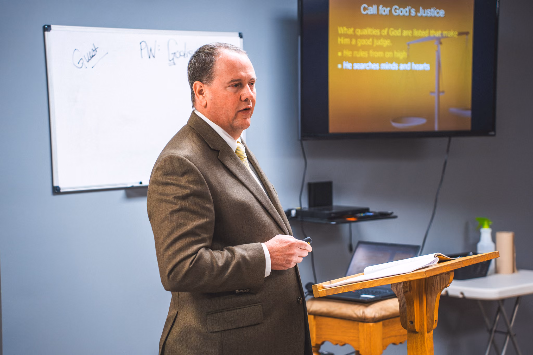 Man in brown suit giving a presentation on God’s justice in a classroom with a podium and whiteboard.