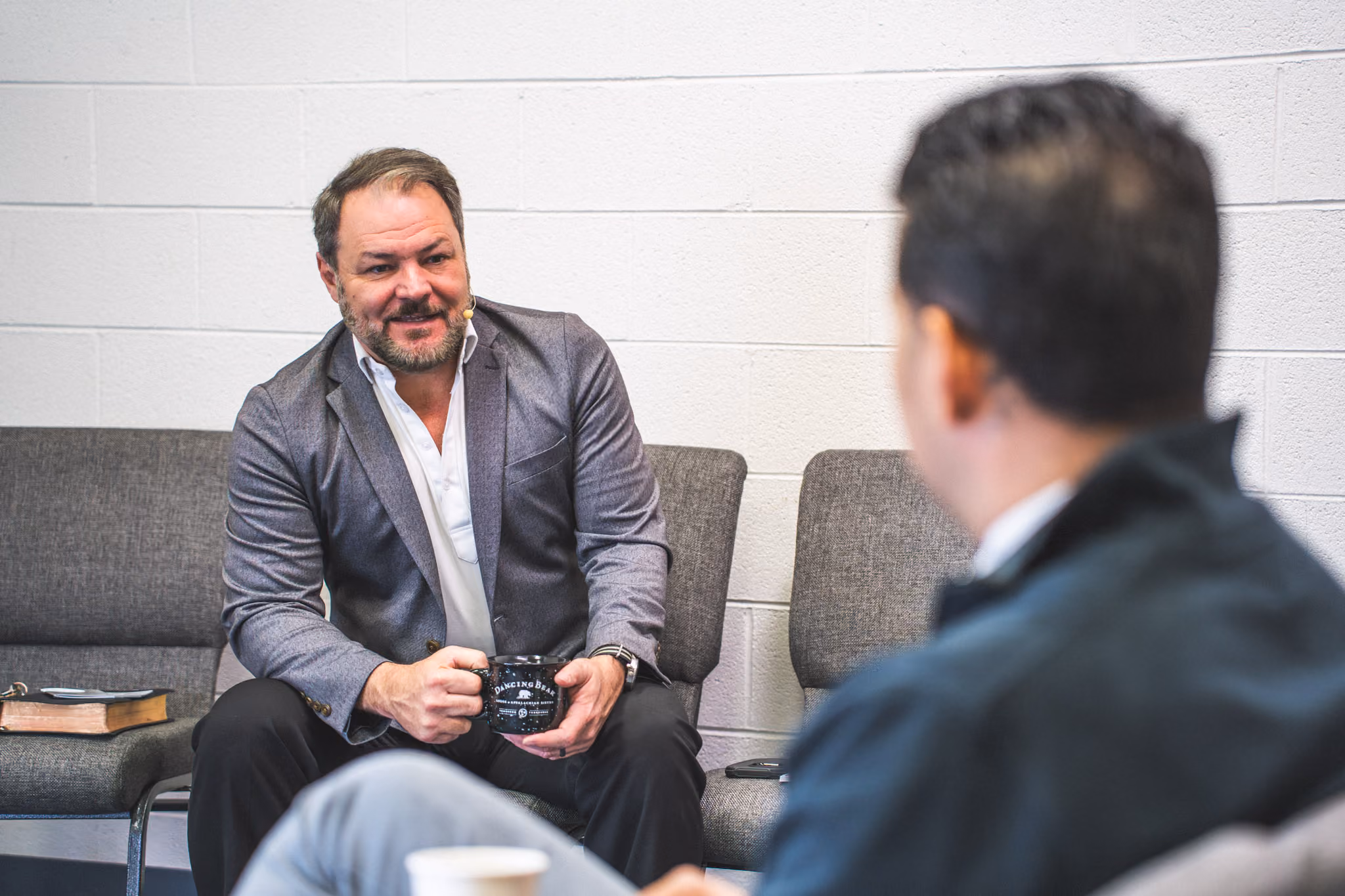 Man in gray blazer holding coffee mug, talking to another man in a casual indoor setting.