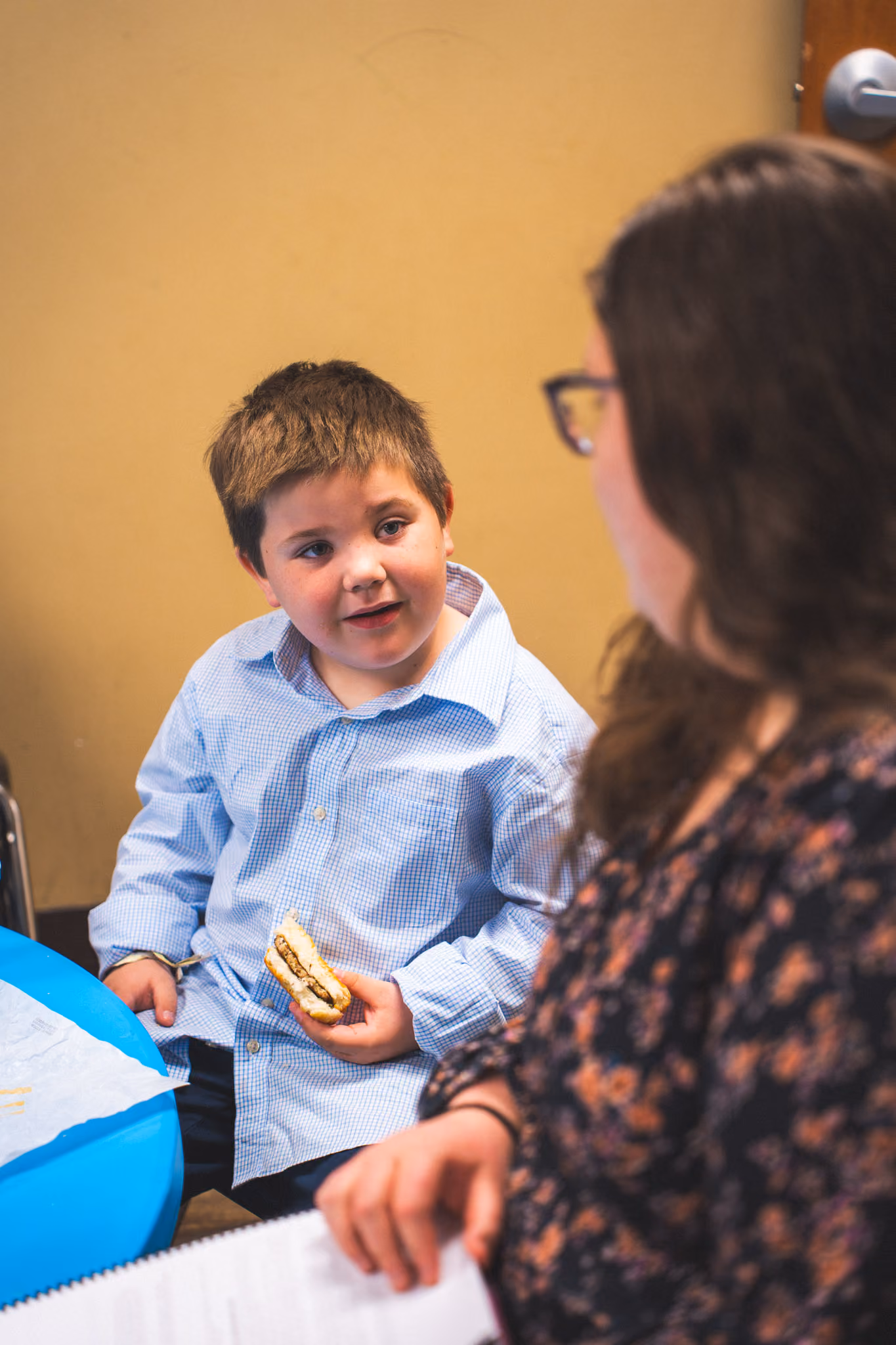 Young boy in a blue shirt eating a sandwich while talking to a woman in glasses.