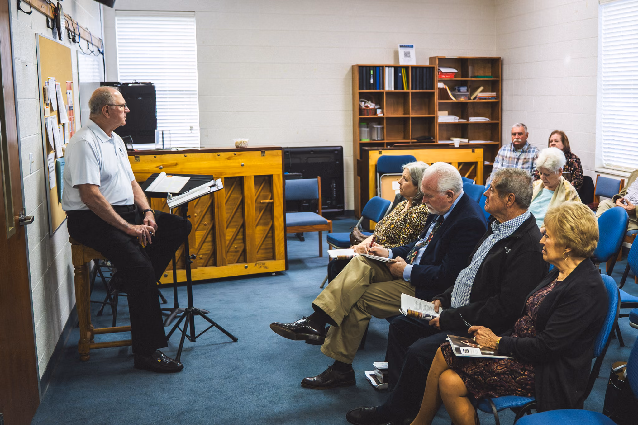Older man speaking to seated audience in small church room with piano and bookshelves.