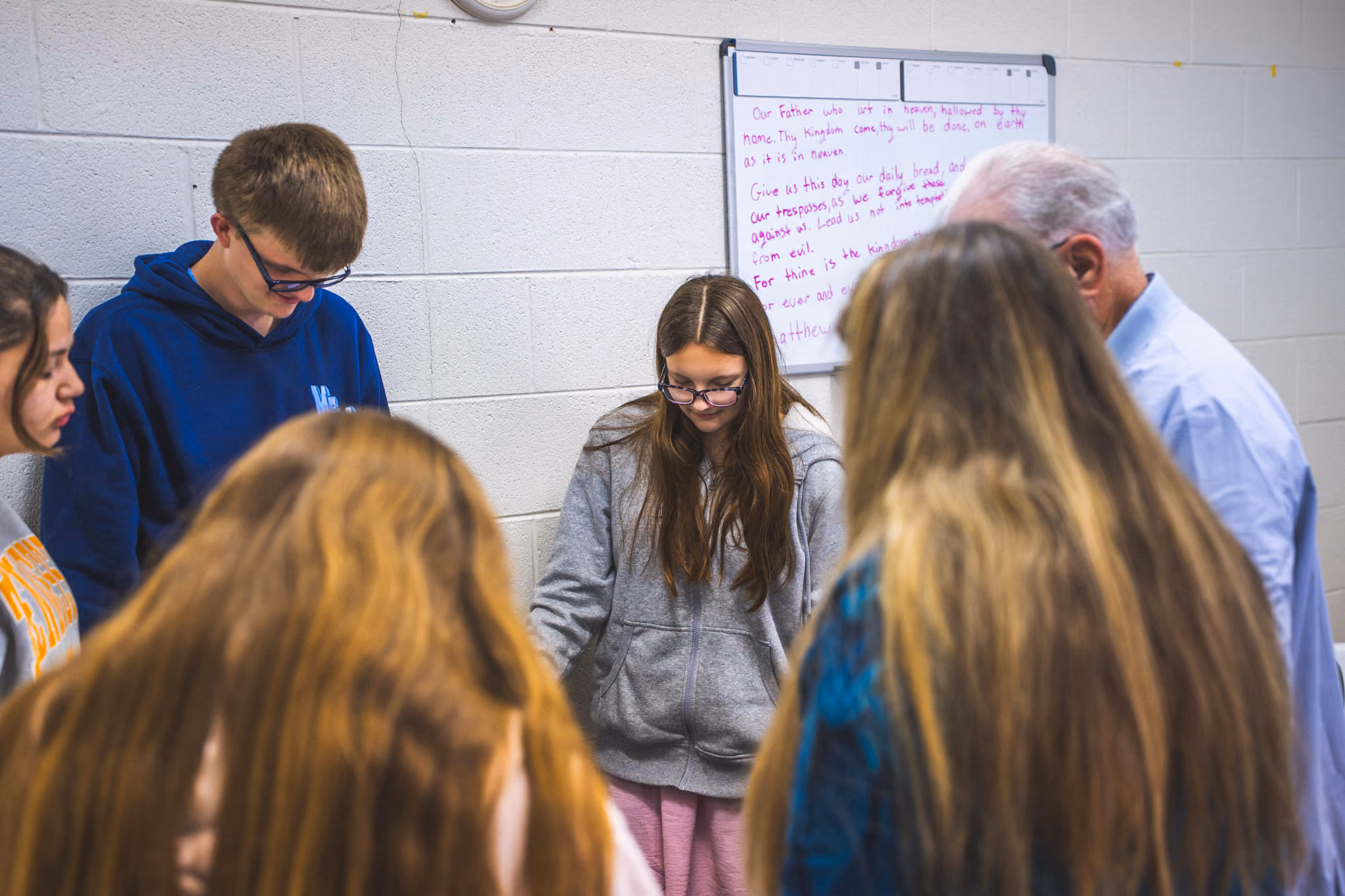 Group of people standing in a circle with heads bowed in prayer in a room with a prayer on a whiteboard.