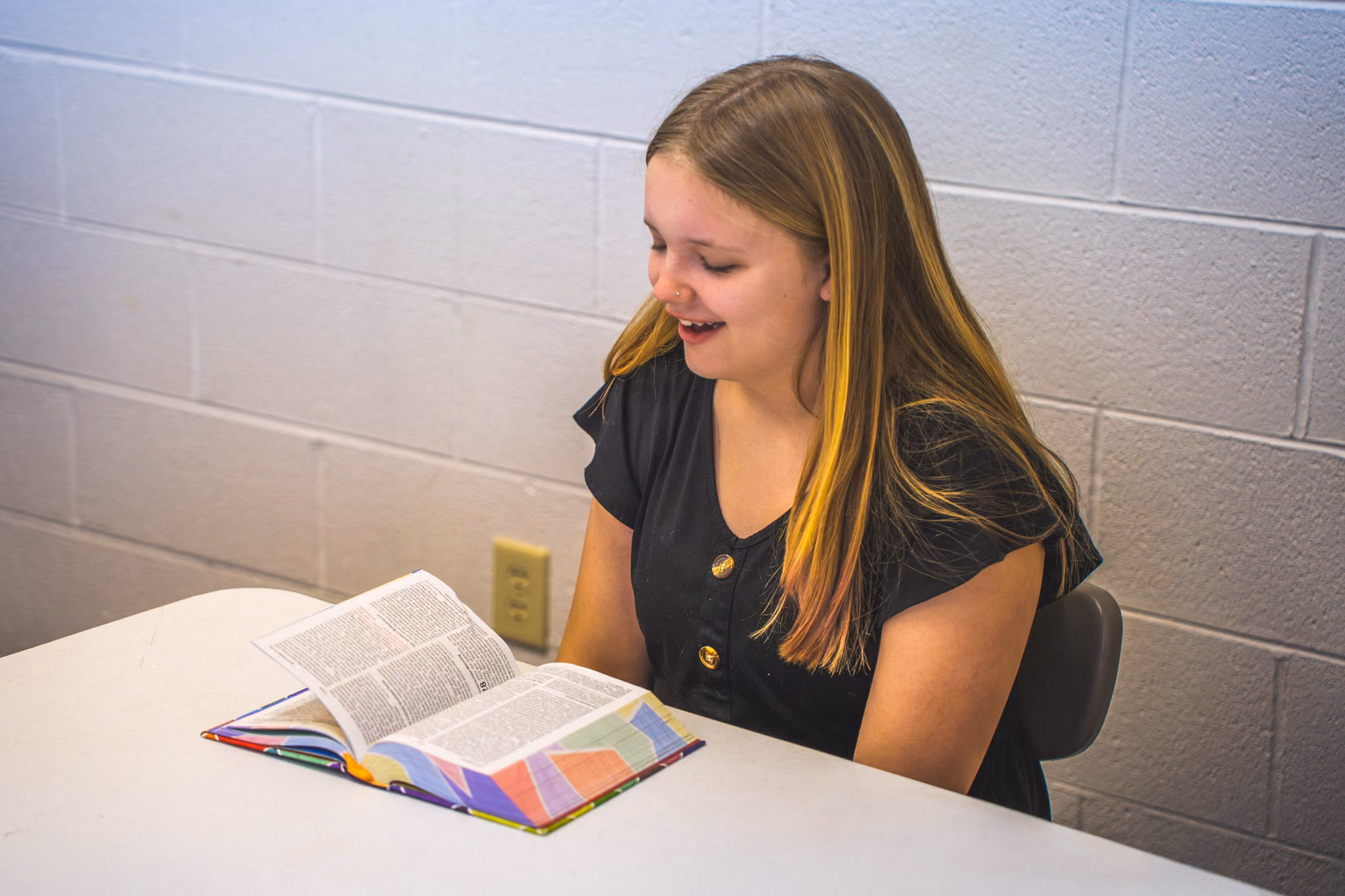 Young woman with long hair reading a colorful book at a white table against a cinder block wall.