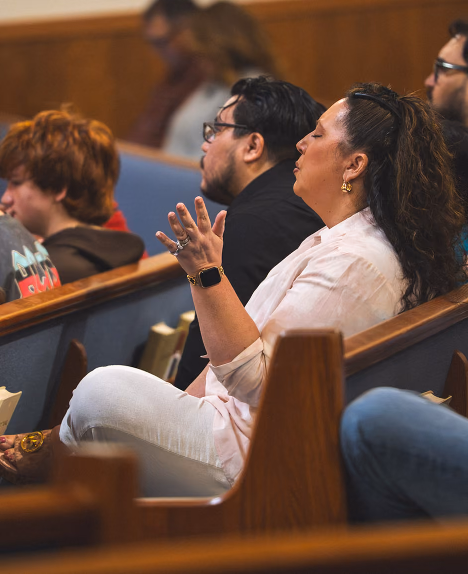 People sitting in pews, woman with eyes closed and hand raised, appearing to pray or worship.