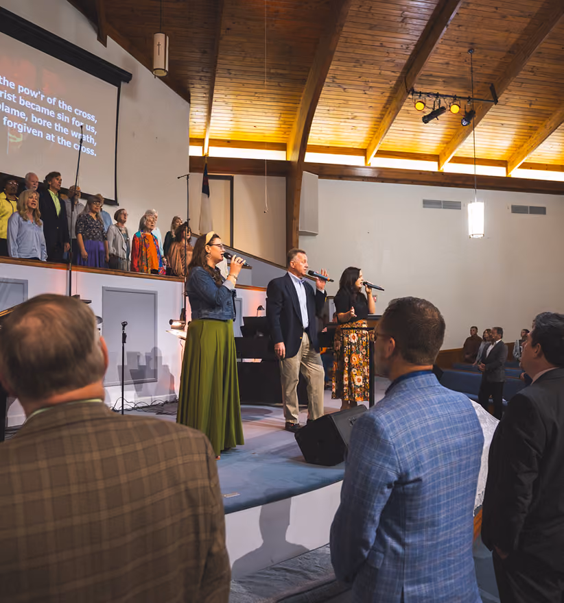 Three people singing on stage with a choir and audience in a church with wooden ceiling beams.
