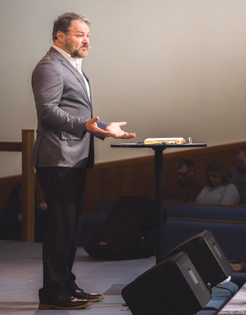 Man in blazer speaking with hands open on a stage near a table with a Bible and glass.
