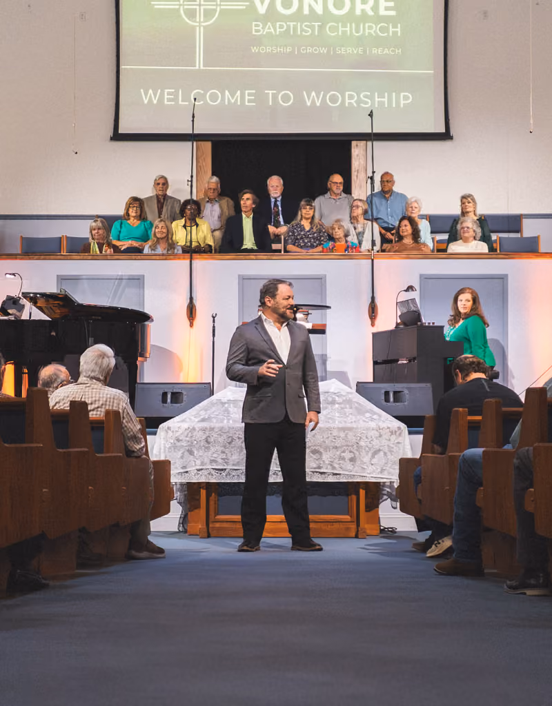 Man speaking in front of church congregation with choir behind and a piano player on stage.