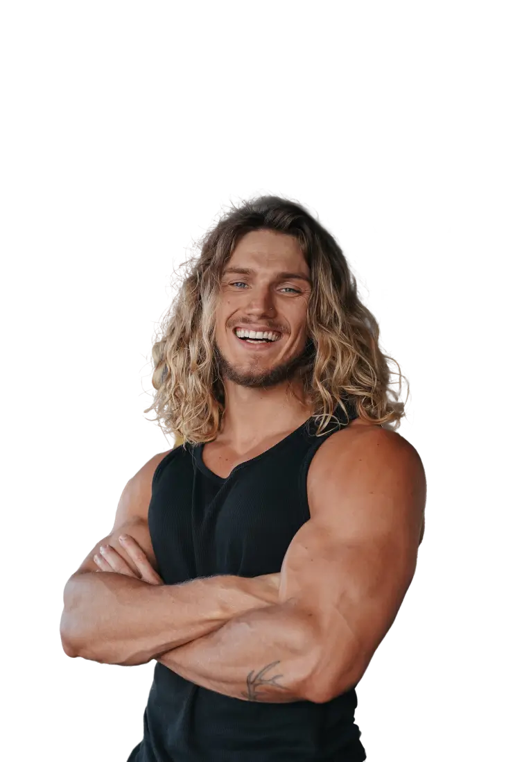 A muscular man with long, wavy hair smiling confidently while crossing his arms. He is wearing a black sleeveless shirt, and the image includes a logo in the top right corner reading 'FITBENY COACHING.
