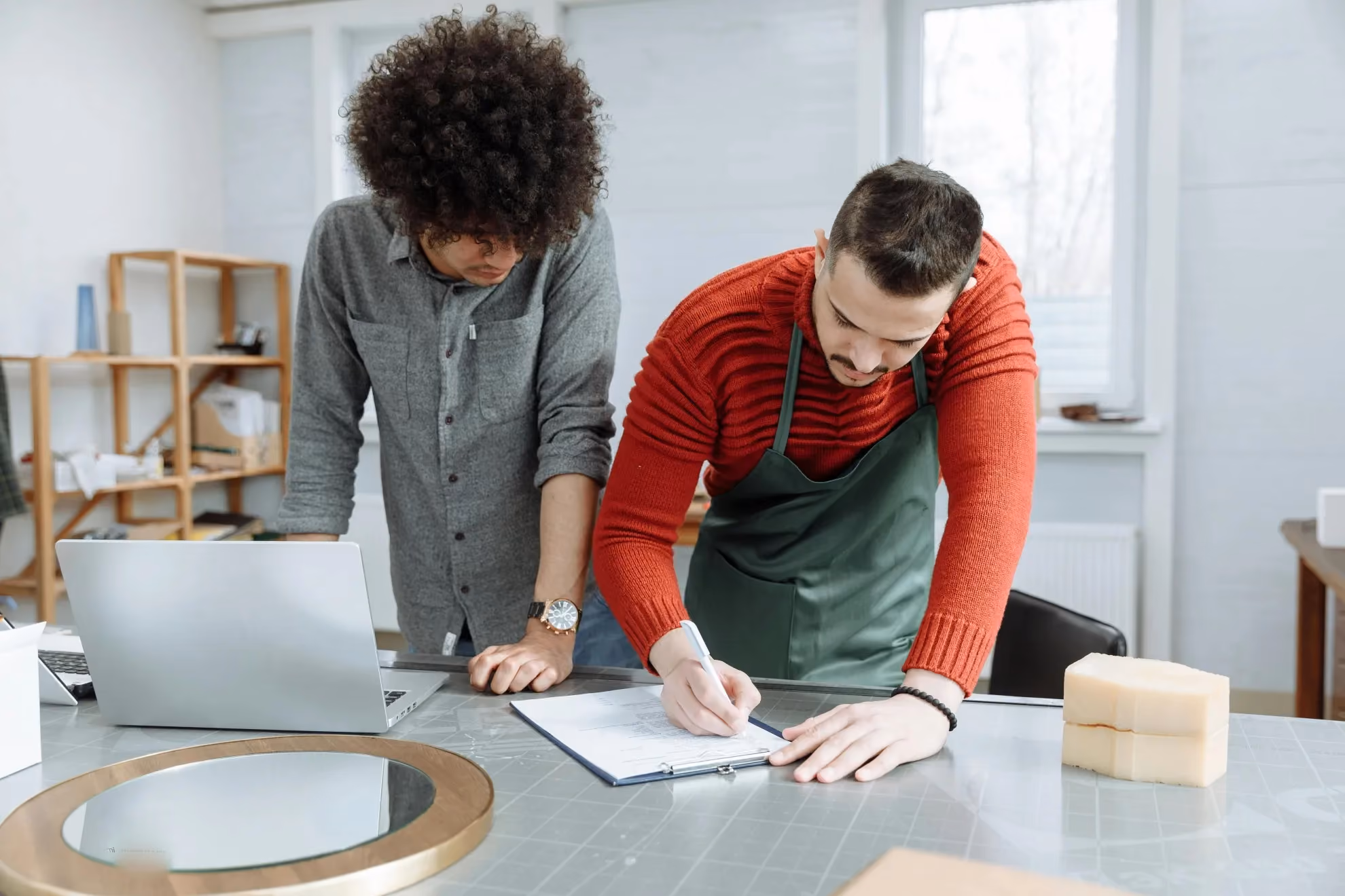 Two men working at a table; one is writing on a clipboard while the other watches, with a laptop and a round mirror nearby.