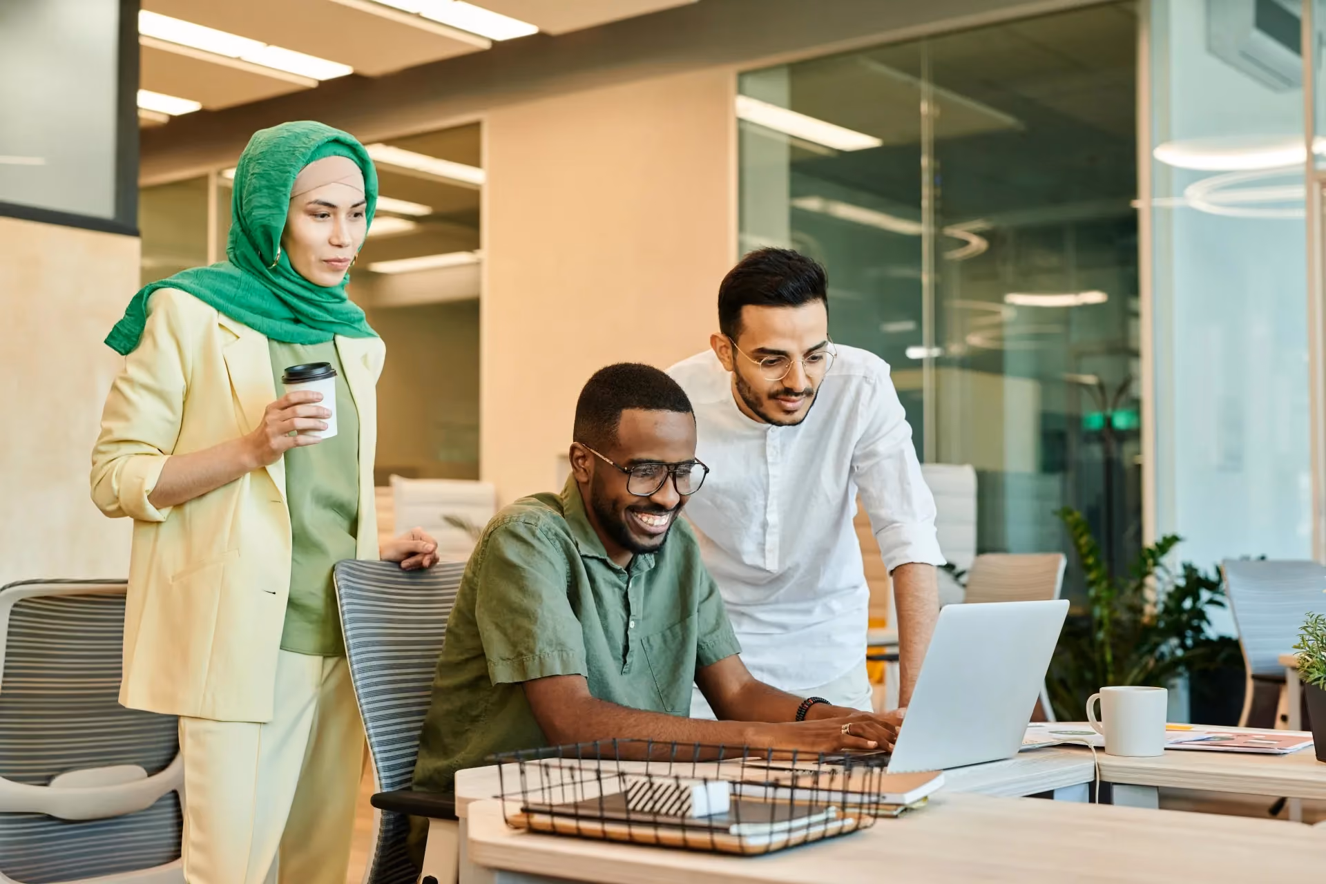 Three diverse coworkers collaborating and smiling while looking at a laptop in a modern office.